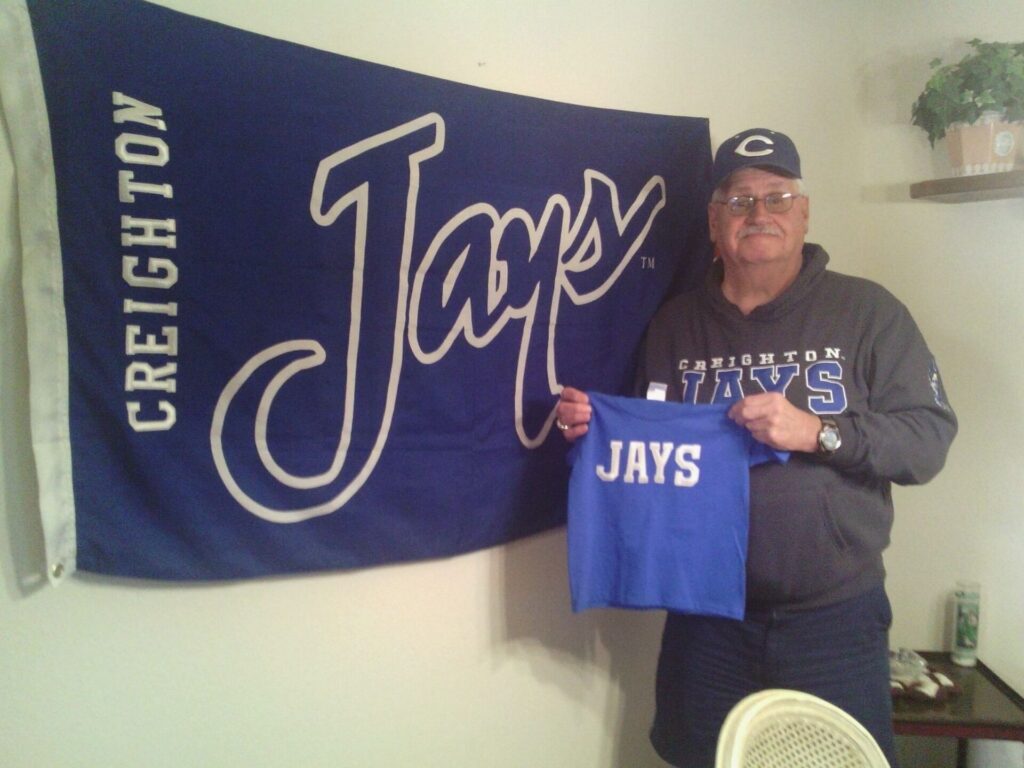 A man, John Bachechi, stands beside a Creighton flag and holds a Creighton shirt. 