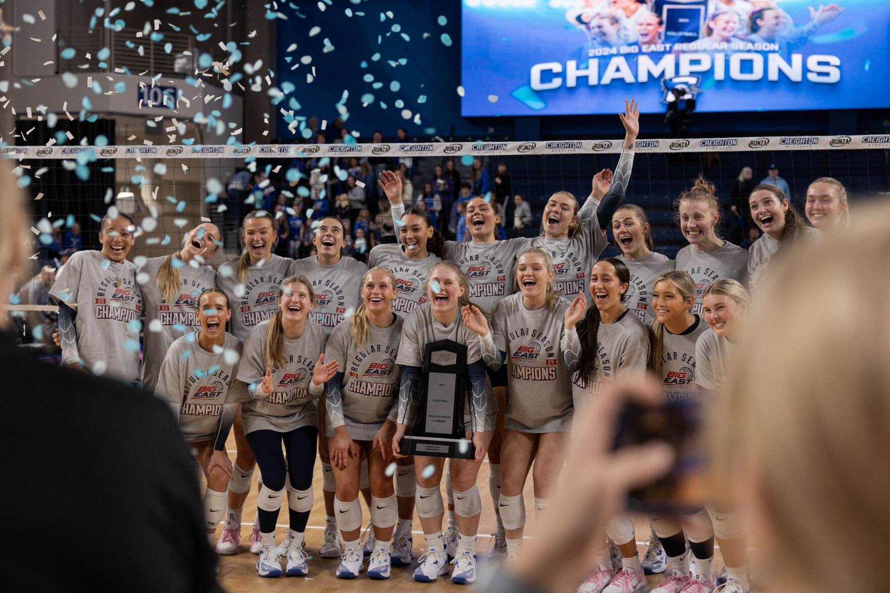 The Creighton volleyball team stands together holding the regular season championship trophy with confetti falling around them.