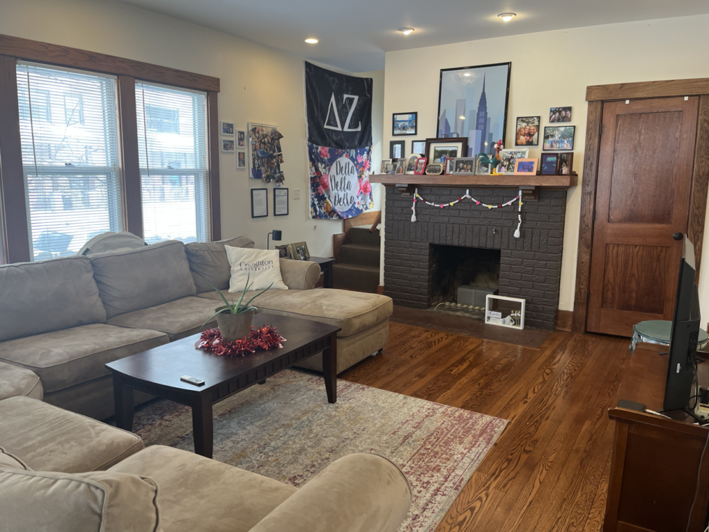 A picture of an open-concept living room featuring a gray L-shaped couch in foreground surrounding a black coffee table set on a rug, with a fireplace in the background adorned with various mantle decorations.